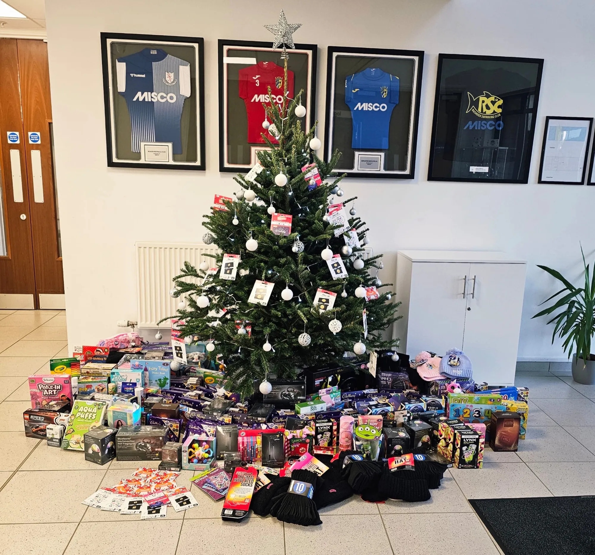 A decorated Christmas tree in an office reception with donated toys, chocolates, thermal socks and hats piled underneath