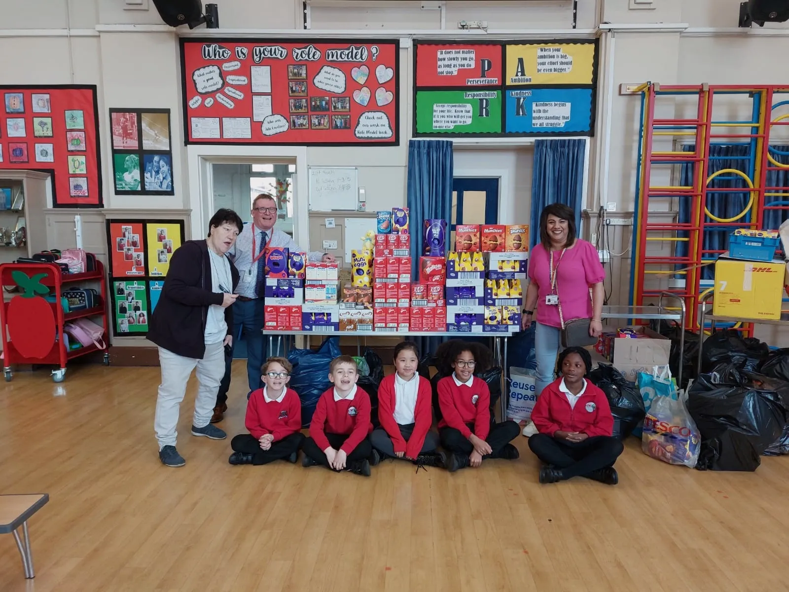 Schoolchildren sitting in a school hall in front of a large stack of donated Easter eggs, with staff standing behind