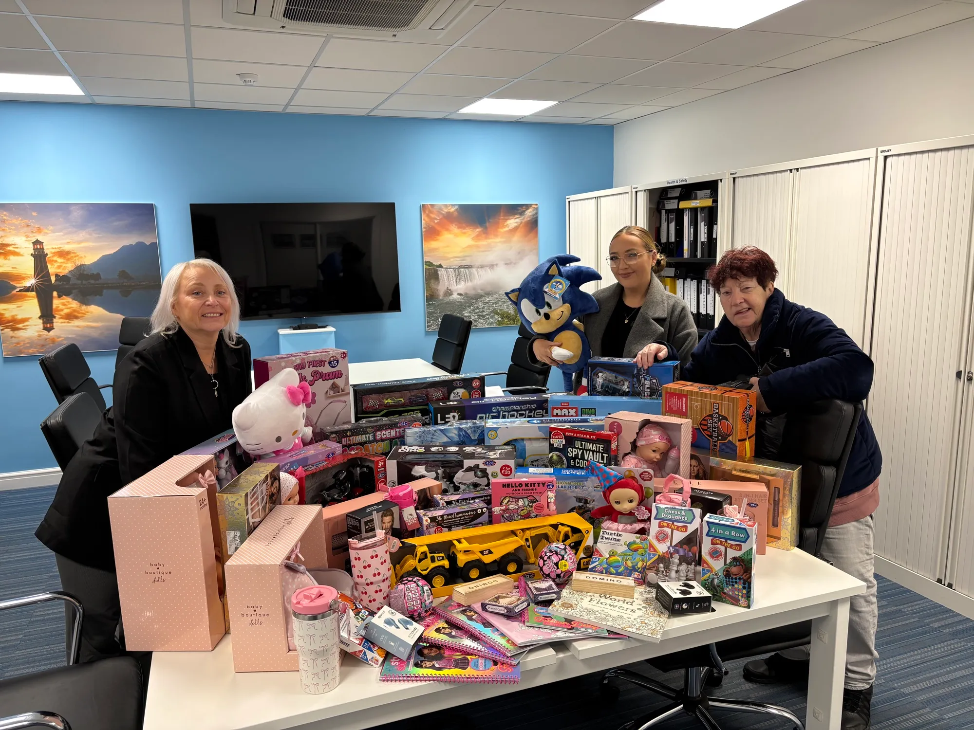 Jeanette Walsh and two workplace volunteers behind a boardroom table covered in donated toys, dolls, games and craft sets