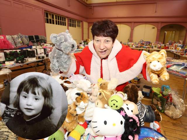 Jeanette Walsh holding up donated teddy bears among piles of soft toys, with an inset black-and-white photograph of herself as a young child